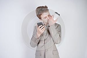 Young teen caucasian student standing in grey business suit, calling by smart phone, drinking coffee, touching his nose and lying