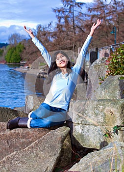 Young teen girl arms raised while sitting on large rock along lake shore, happy