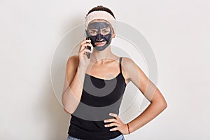 Young teen girl with black clay facial mask making phone call, standing isolated over white background, wearing headband and t