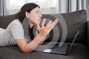 Young teen girl chatting with friends online on laptop lying on sofa