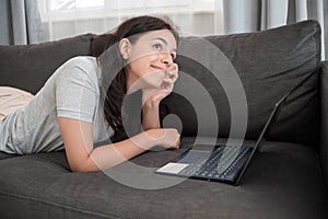 Young teen girl chatting with friends online on laptop lying on sofa