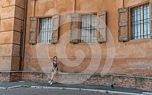 Young teen girl with headband, shorts, tanktop and sneakers sitting on brick bench under 3 windows with shutters and bars