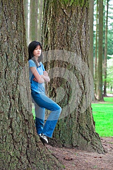 Young teen girl leaning against large pine tree trunk, sad