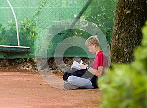 Young teen kid sits on ground under a tree, and reads from folded papers.