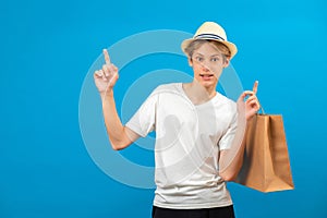 Young teen man points finger aside and holding paper bag in hand isolated on blue. Friendly well-looking boy paying your attention