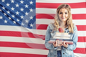 Young teen student holds a stack of study papers and apple on the top with a flag of the Unites States behind her