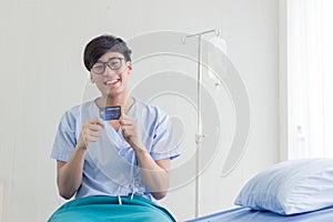 Young teenage Asian man holding credit card sitting on the bed in the hospital