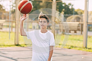 A young teenage basketball player in a white t-shirt stands on the basketball court, twirling a basketball on his