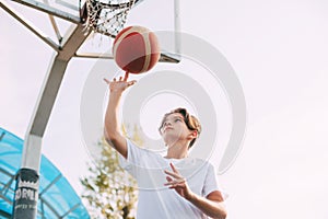 A young teenage basketball player in a white t-shirt stands on the basketball court, twirling a basketball on his