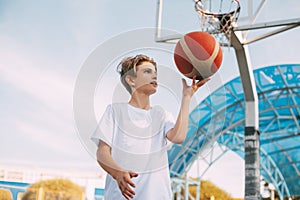 A young teenage basketball player in a white t-shirt stands on the basketball court, twirling a basketball on his