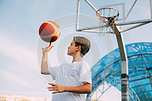 A young teenage basketball player in a white t-shirt stands on the basketball court, twirling a basketball on his