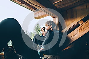 Young teenage boy with hot tea cup drinking while he sitting on the wooden forest house terrace and enjoying the morning spruce