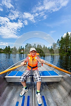 Young teenage boy rowing a rowboat on a lake with blue summer sky in the background.