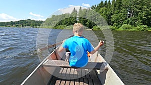 Young teenage boy rowing a rowboat or paddle boat on a lake with forest at bank. Blue summer sky