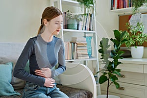 Young teenage female experiencing abdominal pain, sitting on couch at home