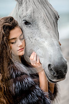 A young teenage girl in a fur coat, with long curly hair in a soft blue dress hugs a white horse on the Bank of the oken