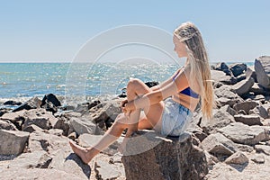 A young teenage girl with long blonde hair blonde sits on the rocks of the breakwater near the sea shore on the beach