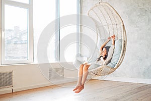 Young teenage girl relaxing in comfortable hanging chair near window at home. Child sitting in chair and chilling out.