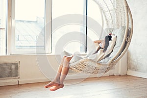 Young teenage girl relaxing in comfortable hanging chair near window at home. Child sitting in chair and chilling out. Relax conce
