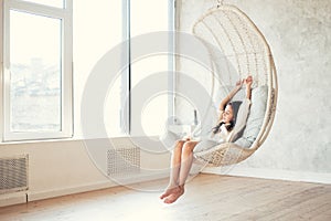 Young teenage girl relaxing in comfortable hanging chair near window at home. Child sitting in chair and chilling out. Relax conce