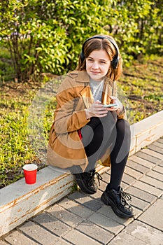 Young teenage girl sits on the street with a smartphone in her hand, headphones, listens to music