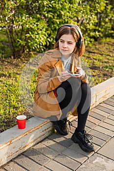 Young teenage girl sits on the street with a smartphone in her hand, headphones, listens to music