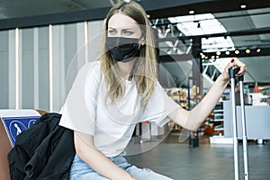 Young teenage girl is sitting at the airport waiting for her flight next to her suitcase. Girl wears a mask