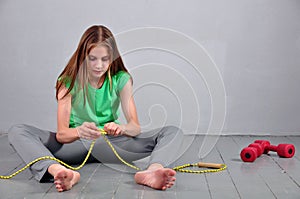 Young teenage girl sitting on the floor with skipping rope and dumbbells relaxing having rest in studio.