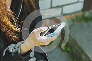 A young teenage girl is sitting outside on the street and holding a cell phone with a blank screen in her hands. Mobile phone in h