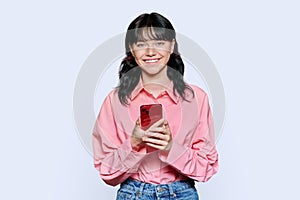 Young teenage girl with smartphone looking at camera on light background