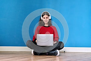 Young teenage guy in headphones with laptop sitting on floor on blue background