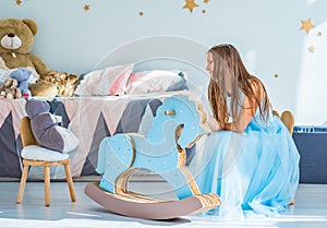 Young teenager brunette girl with long hair in blue dress sitting on chair in her bedroom and play with childrenâ€™s toy