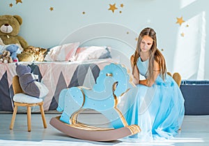 Young teenager brunette girl with long hair in blue dress sitting on chair in her bedroom and play with childrenÃ¢â‚¬â„¢s toy