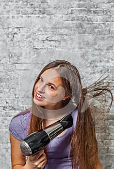 Young teenager brunette girl with long hair dries hair with electrical hair dryer on gray wall background