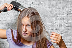 Young teenager brunette girl with long hair dries hair with electrical hair dryer on gray wall background