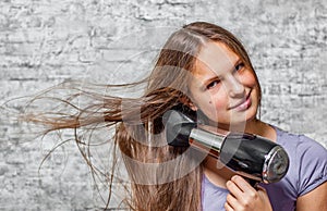 Young teenager brunette girl with long hair dries hair with electrical hair dryer on gray wall background