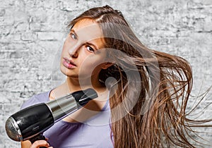 Young teenager brunette girl with long hair dries hair with electrical hair dryer on gray wall background