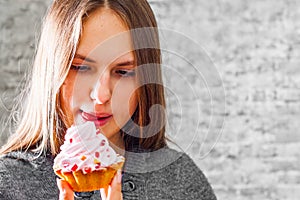 Young teenager brunette girl with long hair eating cream cake on gray wall background