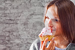 Young teenager brunette girl with long hair eating cream cake on gray wall background