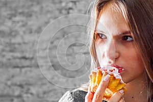 Young teenager brunette girl with long hair eating cream cake on gray wall background