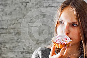 Young teenager brunette girl with long hair eating cream cake on gray wall background