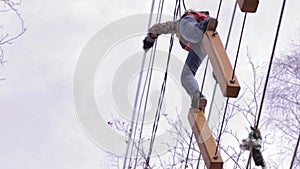 Young teenager climber walks by pendant log bridge on high ropes course in extreme park