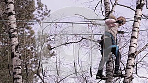 Young teenager climber walks by pendant log bridge on high ropes course in extreme park