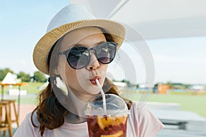 Young teenager girl in hat sunglasses smiling and drinking cool berry cocktail on a hot summer day in outdoor cafe