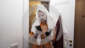 Young teenager girl leaving the bathroom at home with a towel on her head, holding smartphone and hairdryer. Action