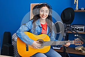 Young teenager girl playing classic guitar at music studio looking positive and happy standing and smiling with a confident smile