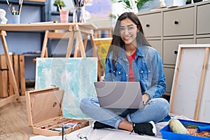 Young teenager girl sitting at art studio using laptop looking positive and happy standing and smiling with a confident smile
