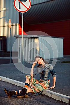 Young teenager girl sitting on the road outside near urban red wall background in skirt and jeans jacket on the sunset.