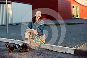 Young teenager girl sitting on the road outside near urban red wall background in skirt and jeans jacket on the sunset.