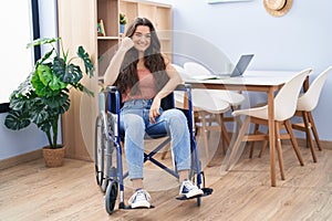 Young teenager girl sitting on wheelchair at the living room smiling happy and positive, thumb up doing excellent and approval